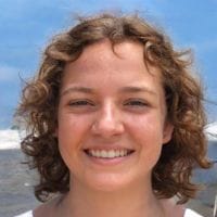 Smiling young person with short curly hair standing outdoors by the ocean under a blue sky