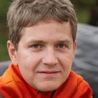Close-up of a smiling young adult with short light brown hair wearing an orange jacket, blurred outdoor background.