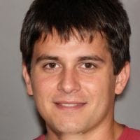 Young man with short dark hair and brown eyes smiling slightly against a plain gray background