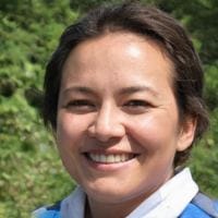 Smiling person with short dark hair wearing a blue jacket standing outdoors with green foliage in the background