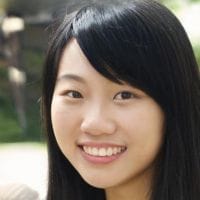 Young Asian woman with long black hair smiling at the camera, outdoor blurred greenery in background.