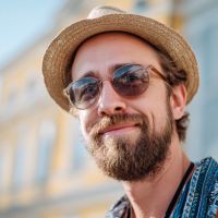 Smiling bearded man wearing round sunglasses and a straw hat, with a patterned shirt and blurred building in the background.