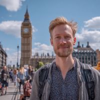 Smiling man with short blond hair and backpack standing on Westminster Bridge with Big Ben and Houses of Parliament in the background on a sunny day