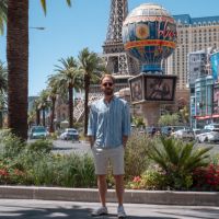 Man wearing sunglasses, striped short-sleeve shirt and shorts standing on a Las Vegas street with palm trees and the Paris hotel’s replica Eiffel Tower and hot-air-balloon sign in the background.