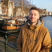 Young man wearing a tan winter jacket standing by a canal with moored boats and buildings in soft sunlight.