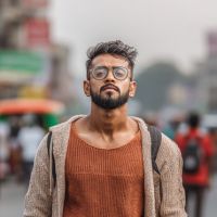 Young man with glasses, short curly hair and a beard wearing a rust-colored knit sweater and beige cardigan, standing on a busy urban street.