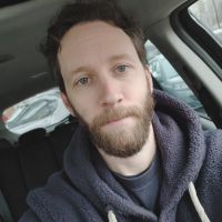 Bearded man wearing a dark hooded sweater sitting in a parked car and looking toward the camera.